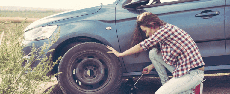 Young woman fixing a flat tire on her car during a road trip, showing budget travel challenges.