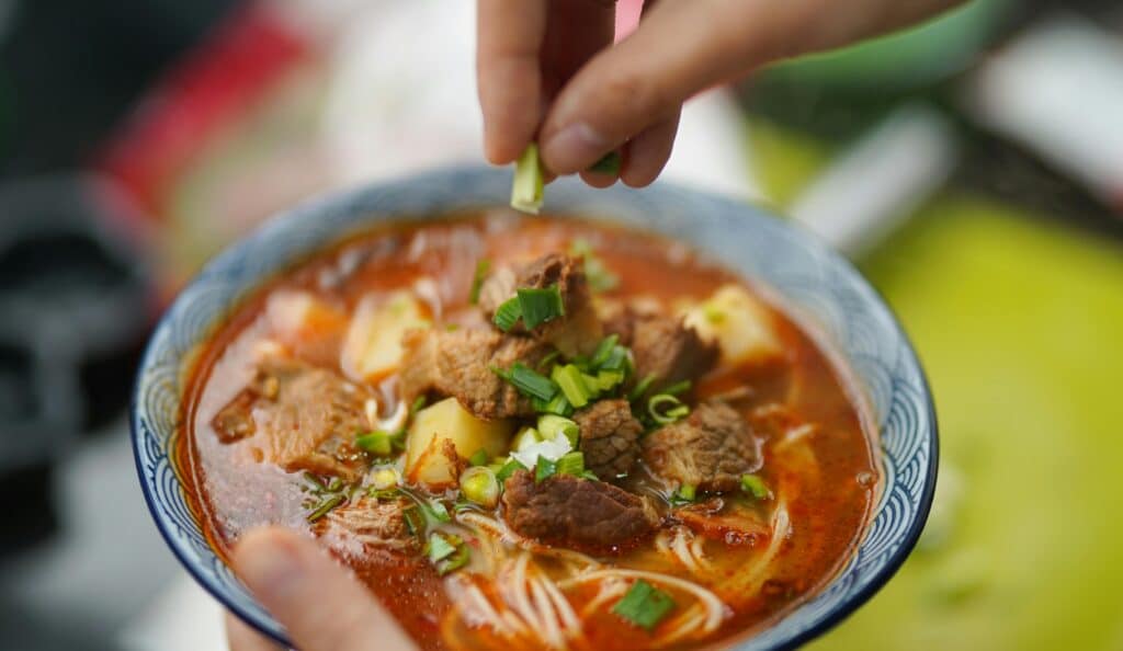 Close-up of a bowl of steaming beef noodle soup garnished with fresh spring onions at a night market